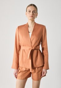 Peach satin blazer with a shawl collar and a tied waist. Long sleeves and a structured fit. Paired with matching shorts.