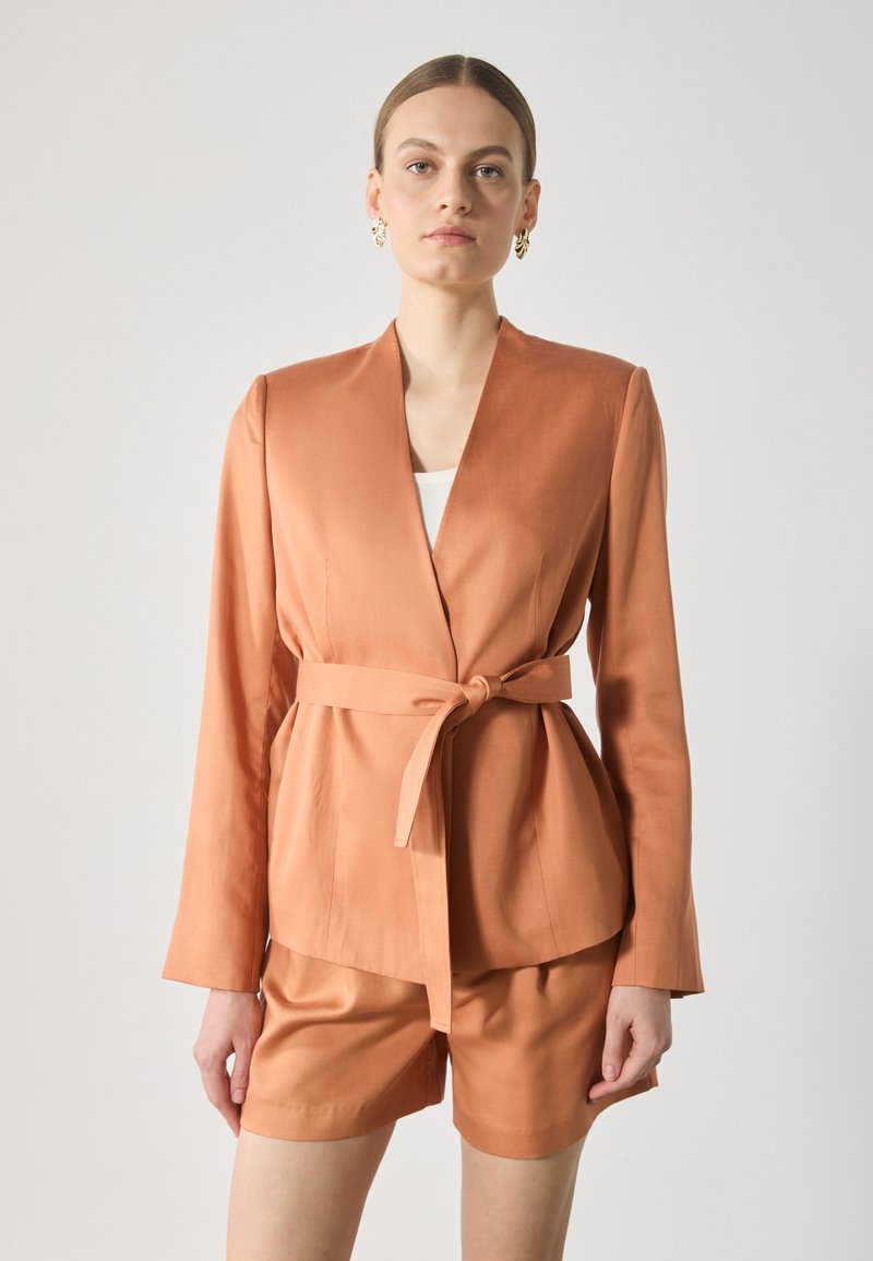 Peach satin blazer with a shawl collar and a tied waist. Long sleeves and a structured fit. Paired with matching shorts.