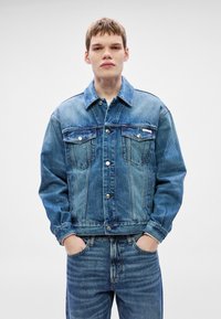 Τζιν μπουφάν μέσης μπλε απόχρωσης με κλασικό γιακά, κούμπωμα με κουμπιά, δύο τσέπες στο στήθος και αντίθετη ραφή· φορεμένο με τζιν παντ�ελόνι στο ίδιο χρώμα.