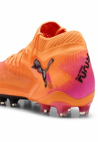 Kirkkaan oranssi ja vaaleanpunainen Puma-jalkapallokenkä, jossa musta logo, teksturoitu neulepinta ja niiteillä varustettu pohja koville kentille.