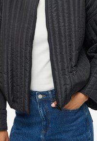 Juodas siuvinėtas švarkas su užtrauktuku, turintis raukšlėtą tekstūrą ir apvalų apykaklę, apsivilktas ant baltos palaidinės ir mėlynų džinsinių sijonų.