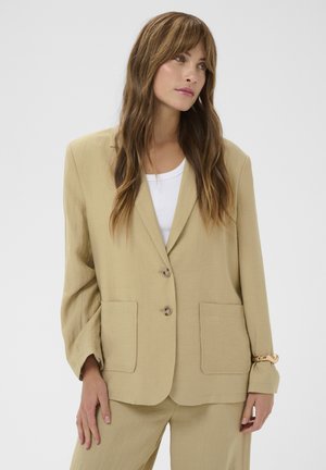 Vrouw met lang bruin haar, gekleed in een beige colbert over een wit shirt, staand met een hand in de zak, kijkend naar rechts, neutrale achtergrond.