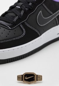 Svart Nike-sneaker med texturerade överlägg, vit mellansula och lila detaljer. Framifrån syns en guld- och svart Nike-emblem med detaljerad gravyr.