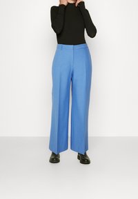 Světle modré široké kalhoty vyrobené z hladké tkaniny, se zvýšeným pasem a čistými puky, v kombinaci s přiléhavým černým topem a černými botami.