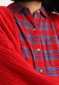 Rode geribbelde cardigan over een rode en blauwe geruite overhemden met knopen. Beschikt over knoopsluitingen en een geklede kraag.