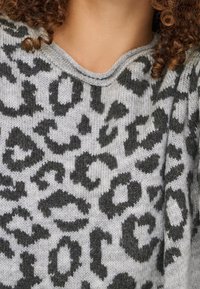 Personne aux cheveux bouclés portant un pull en maille gris avec un motif léopard noir et gris foncé et un col rond.
