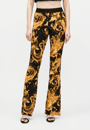 Ženska v črnih razširjenih hlačah z drzno zlato baročnimi vzorci in črnih odprtih petah, stoji na preprostem ozadju.
