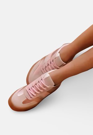 Jambes portant des baskets roses brillantes avec des lacets rose clair, reposant sur un fond blanc uni.