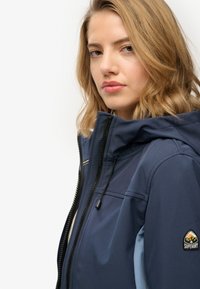 Granatowa, wodoodporna kurtka z kapturem, zamkiem błyskawicznym i naszywką z logo na rękawie. Gładka tekstura z dwukolorowym wzorem.