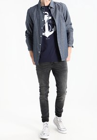 Camisa de mezclilla de manga larga, color azul, con botones en la parte delantera sobre una camiseta gráfica azul marino con un diseño de ancla, combinada con jeans oscuros ajustados y zapatillas blancas.