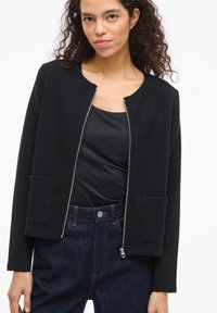 Μαύρο μπουφάν με φερμουάρ, με υφή, στρογγυλή λαιμόκοψη και δύο μπροστινές τσέπες. Φοράται πάνω από μαύρη μπλούζα και συνδυάζεται με σκούρο τζιν.