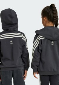 Felpa grigia con cappuccio, caratterizzata da strisce bianche sulle spalle, con il logo Adidas e Disney sulla schiena e orli e polsini a coste.