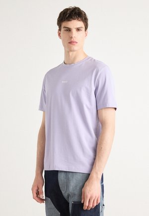 T-shirt en coton lilas à col rond et manches courtes, avec un petit logo blanc sur la poitrine. La coupe est décontractée, associée à un pantalon en denim.