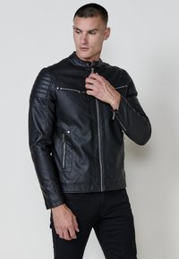 Homme en veste en cuir noir avec des épaules matelassées, des zippers sur la poitrine et les hanches, ajustant le col de sa veste devant un fond uni.