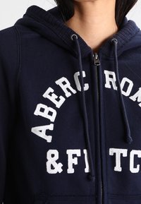 Sudadera con cremallera de color azul marino hecha de una suave mezcla de algodón, con el texto bordado en blanco "ABERCROMBIE & FITCH" y cordones ajustables.