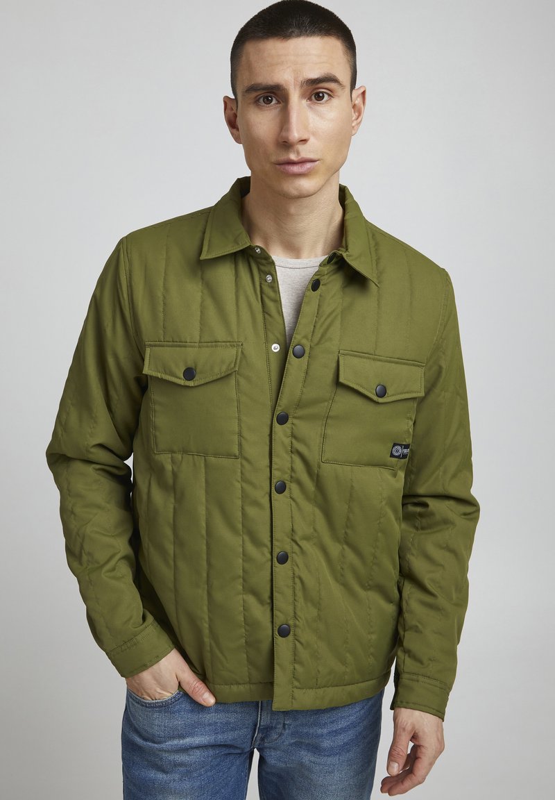 Giubbotto imbottito verde con due tasche sul petto, bottoni a pressione e un orlo leggermente arrotondato. Indossato sopra una camicia grigia chiara.