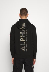 Sudadera negra con un logo de camuflaje "ALPHA INDUSTRIES" impreso verticalmente en la parte trasera, con capucha ajustable y puños acanalados.