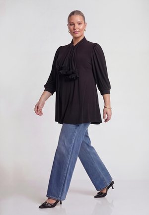 Kvinde med bundet hår bærer sort bluse med flæsedetalje, blå vide jeans, sorte spidse hæle og gyldne hoop-øreringe.