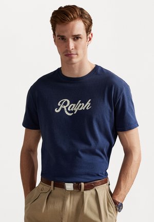 Marineblauwe katoenen T-shirt met "Ralph" in witte cursieve letters, gecombineerd met een beige broek en een bruine riem, met een klassieke ronde hals.