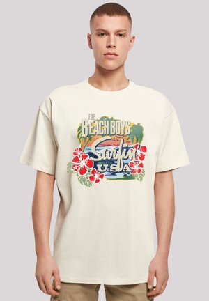 Young man wearing an off-white T-shirt with a colorful "The Beach Boys Surfin' USA" graphic featuring palm trees and red hibiscus flowers.