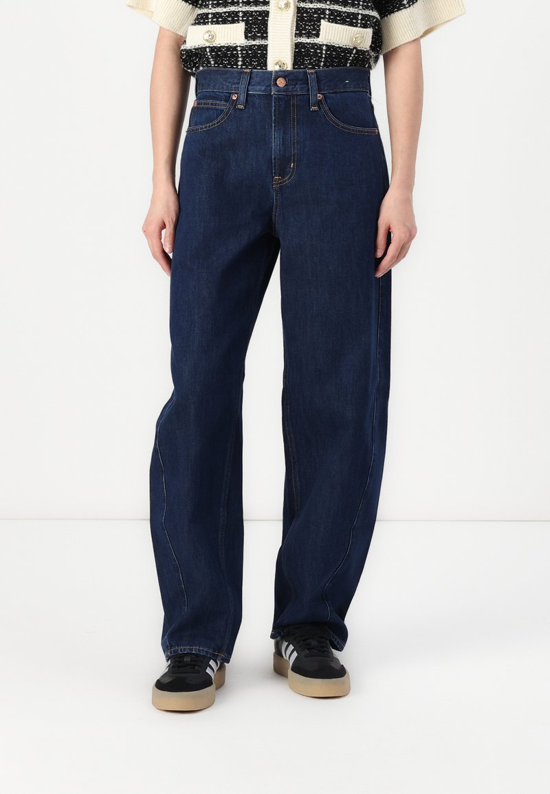Dunkelblaue Weitbeinjeans aus Denim, mit fünf Taschen, Steppnähten und einem hochgeschnittenen Design. Kombiniert mit schwarzen Sneakers.