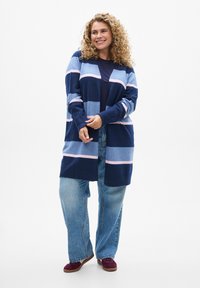 Gilet rayé en bleu, bleu clair et rose ; en tissu tricoté doux, à manches longues, ouvert à l'avant, associé à un jean en denim ample et des chaussures bordeaux.