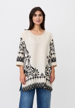 Mujer con una túnica crema con patrones florales negros en las mangas y el dobladillo, combinada con vaqueros azules, de pie frente a un fondo liso y claro.