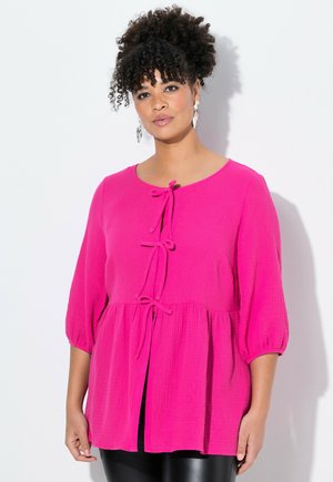 Haut texturé rose avec un col rond, des manches trois-quarts et un détail de nouage à l'avant. Il présente un style peplum et un tissu léger.