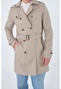 Beige Doppelreiher-Trenchcoat mit Schulterklappen, braunen Knöpfen, Gürtel und seitlichen Taschen, getragen über einem weißen Hemd und blauen Jeans.