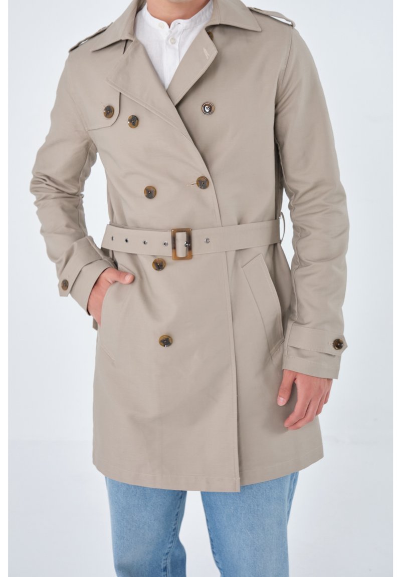 Beige Doppelreiher-Trenchcoat mit Schulterklappen, braunen Knöpfen, Gürtel und seitlichen Taschen, getragen über einem weißen Hemd und blauen Jeans.