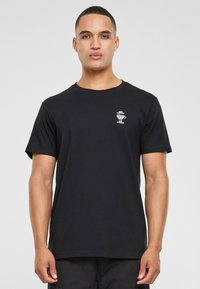 Sort bomulds T-shirt med korte ærmer. Har et lille hvidt grafisk print på brystet. Klassisk rund halsudskæring. Løs pasform.