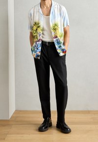 Camisa de manga corta, de color azul claro, con estampado tropical que presenta palmeras y gráficos coloridos, combinada con pantalones negros de corte entallado y zapatos negros.