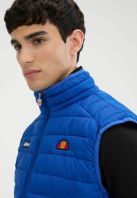 Niebieska pikowana kamizelka bez rękawów z wysokim kołnierzem, zamek błyskawiczny oraz pomarańczowym i żółtym haftowanym logo po lewej stronie klatki piersiowej. Teksturowana powierzchnia tkaniny.