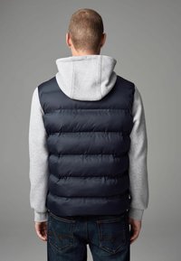 Gilet matelassé bleu marine sur un sweat à capuche gris clair, présentant des sections matelassées horizontales. Le gilet a une texture lisse et un col montant. Un jean en denim foncé est visible.