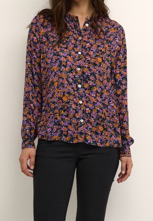 Kobieta ubrana w długą koszulę na guziki z pomarańczowo-fioletowym kwiatowym wzorem, w połączeniu z czarnymi spodniami, stojąca na jednolitym jasnym tle.