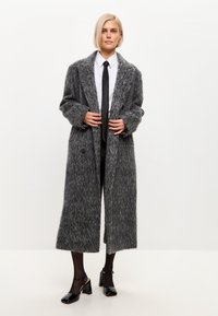 Abrigo largo de punto gris con un acabado texturizado, con un cierre de doble botonadura en el frente, amplios solapas y dos botones; combinado con un atuendo formal y zapatos negros.