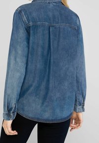 Denimová košile v bledě modré barvě s límcem na knoflíky, dlouhými rukávy a sklady na zadním dílu; má knoflíkové manžety.