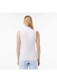 Polo senza maniche di colore bianco, realizzato in tessuto liscio, con un colletto alto e una vestibilità aderente. Indossato con pantaloni azzurri chiari, con un branding minimo visibile.