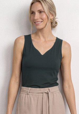 Femme blonde portant un top sans manches vert foncé à col en V et un pantalon beige taille haute avec une ceinture nouée, souriante et regardant vers la droite.