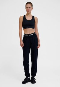 Zwarte sportbeha met een platte band en een wit logo; zwarte joggers met elastische tailleband en logo; gedragen met zwarte sportschoenen.