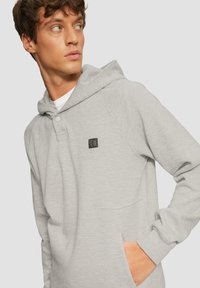 Sudadera gris claro hecha de material suave, con un bolsillo frontal, capucha con cordón y un pequeño parche de logo negro en el pecho.