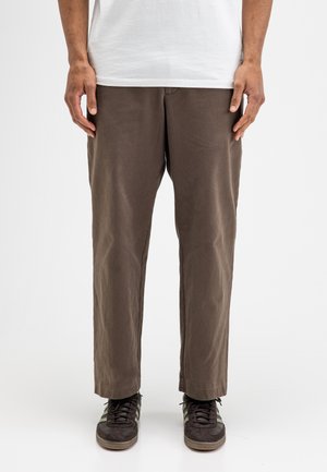 Mann, der braune gerade geschnittene Hosen, schwarz-grüne Sneakers und ein weißes Hemd trägt, vor einem schlichten weißen Hintergrund stehend.
