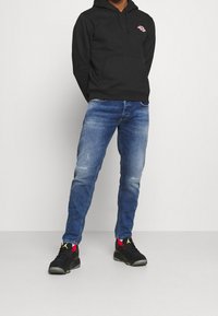 Svart huva med ficka fram och röd logga, tillsammans med medelblå jeans som har blekningar, och svarta sneakers med röda detaljer.