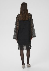 Melnas mežģīņu kleitas, kas izceļas ar slāņainām frizēm, garām piedurknēm un vaļīgu piegriezumu. Apvienota ar caurspīdīgām melnām zeķubiksēm un gaišu nokrāsu kurpēm.