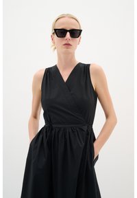 Schwarzes ärmelloses Wickelkleid mit taillierter Taille und seitlichen Taschen. Der Stoff wirkt glatt mit mattem Finish. Model trägt geometrische schwarze Sonnenbrille.