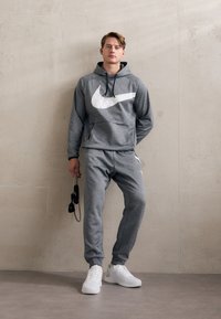 Felpa grigia con grande logo Nike bianco, abbinata a pantaloni della tuta coordinati. Indossata con sneakers bianche, in piedi su un pavimento liscio contro un muro texturizzato.