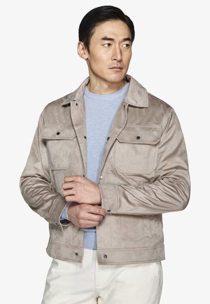 Mężczyzna w jasnoszarej zamszowej kurtce na niebieskim swetrze i białych spodniach, patrzący w bok podczas zapinania mankietu kurtki.