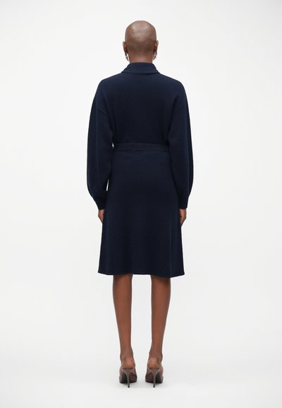 Vestido tricotado azul marinho com gola, corte relaxado, mangas longas e cintura cingida, terminando logo acima dos joelhos. Textura suave.
