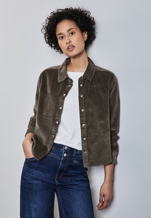 Giacca camicia in corda verde oliva con colletto, chiusura a bottoni e tessuto strutturato, abbinata a jeans in denim blu.