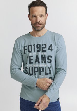 Lichtblauw lang mouw T-shirt met vetgedrukte zwart-witte tekst; zachte stof, ronde halslijn en slimfit ontwerp, gecombineerd met donkere jeans.